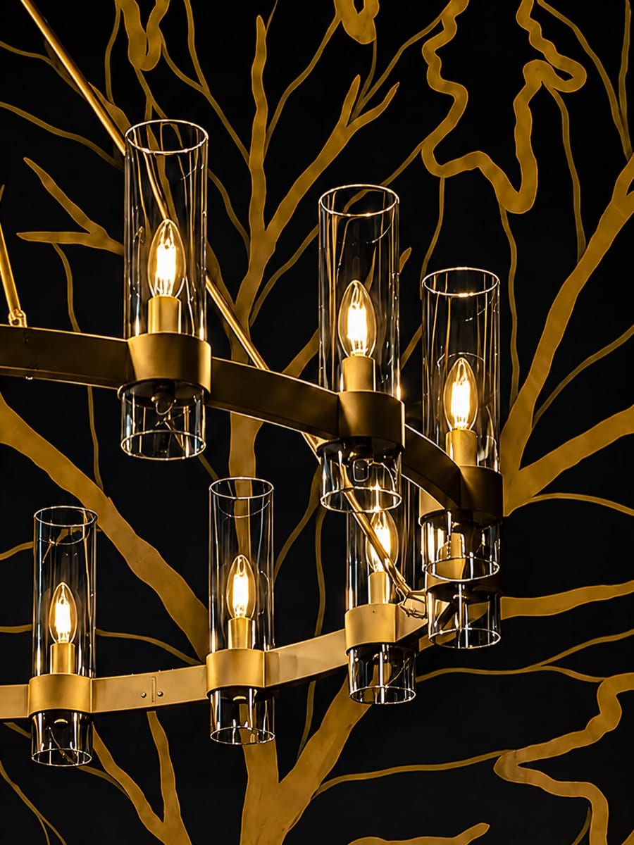 A tiered brass chandelier with glass cylinder shades and warm filament bulbs, hanging in front of a wall painted with a large gold oak-branch mural on black.