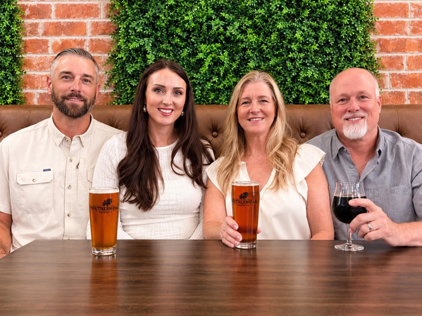 The four owners of Southern Oak Brewing seated side by side in a brown leather booth in front of a brick wall and green hedge panel, three holding pints of amber beer in Southern Oak branded glasses and one holding a glass of dark beer.