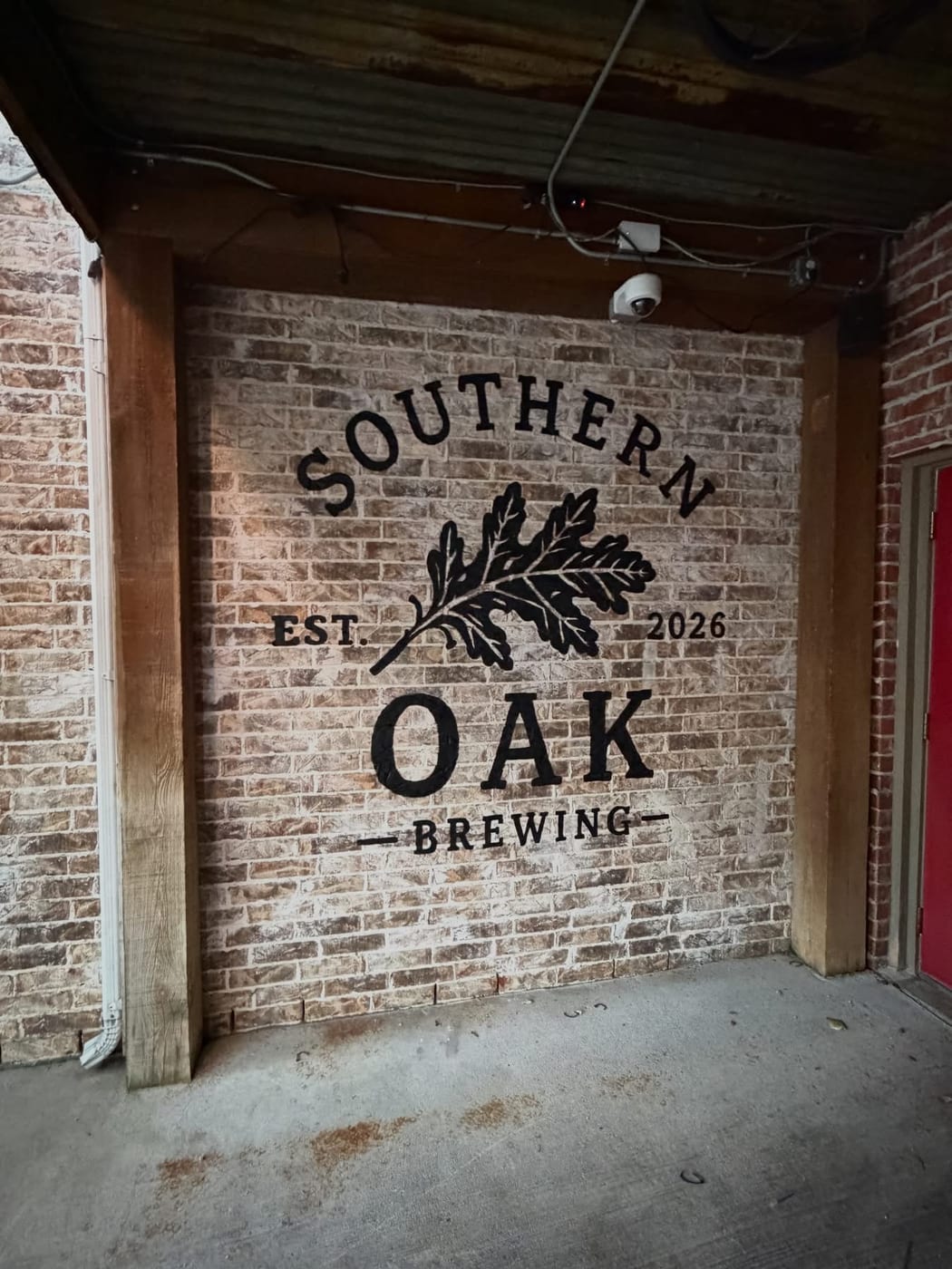 The Southern Oak Brewing logo painted in black on a whitewashed brick wall under the patio's wooden beams, reading SOUTHERN OAK BREWING, EST. 2026, around an oak-leaf mark.